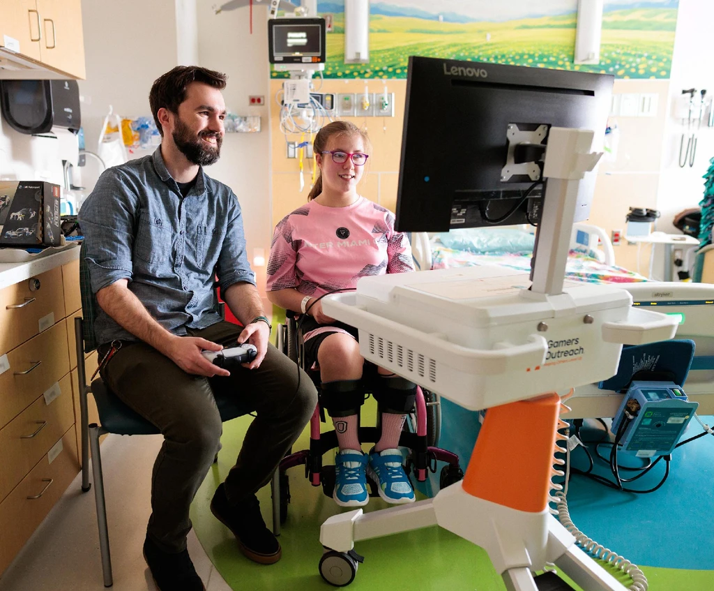 A Gamers Outreach volunteer and a teenage girl in a wheelchair smiling while playing video games together in a hospital room
